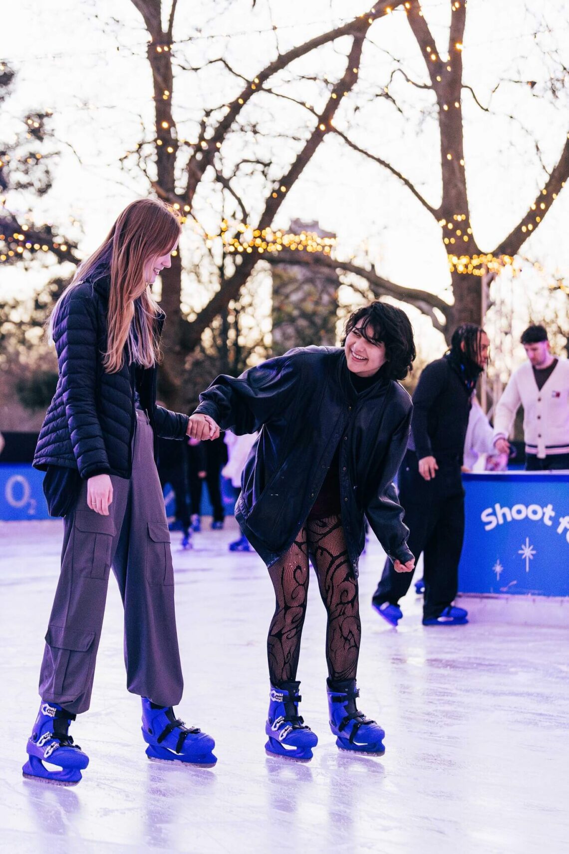 Two young women ice skating outdoors on a rink. One woman wears a black puffer jacket and loose gray pants, while the other is dressed in a black oversized jacket and patterned tights. In the background, people skate and fairy lights are visible on trees.