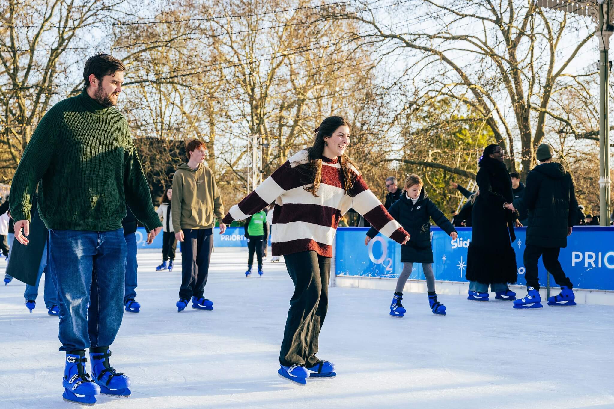 People ice skating outdoors on a sunny day, wearing winter clothing and blue rental skates, with trees and other skaters in the background.