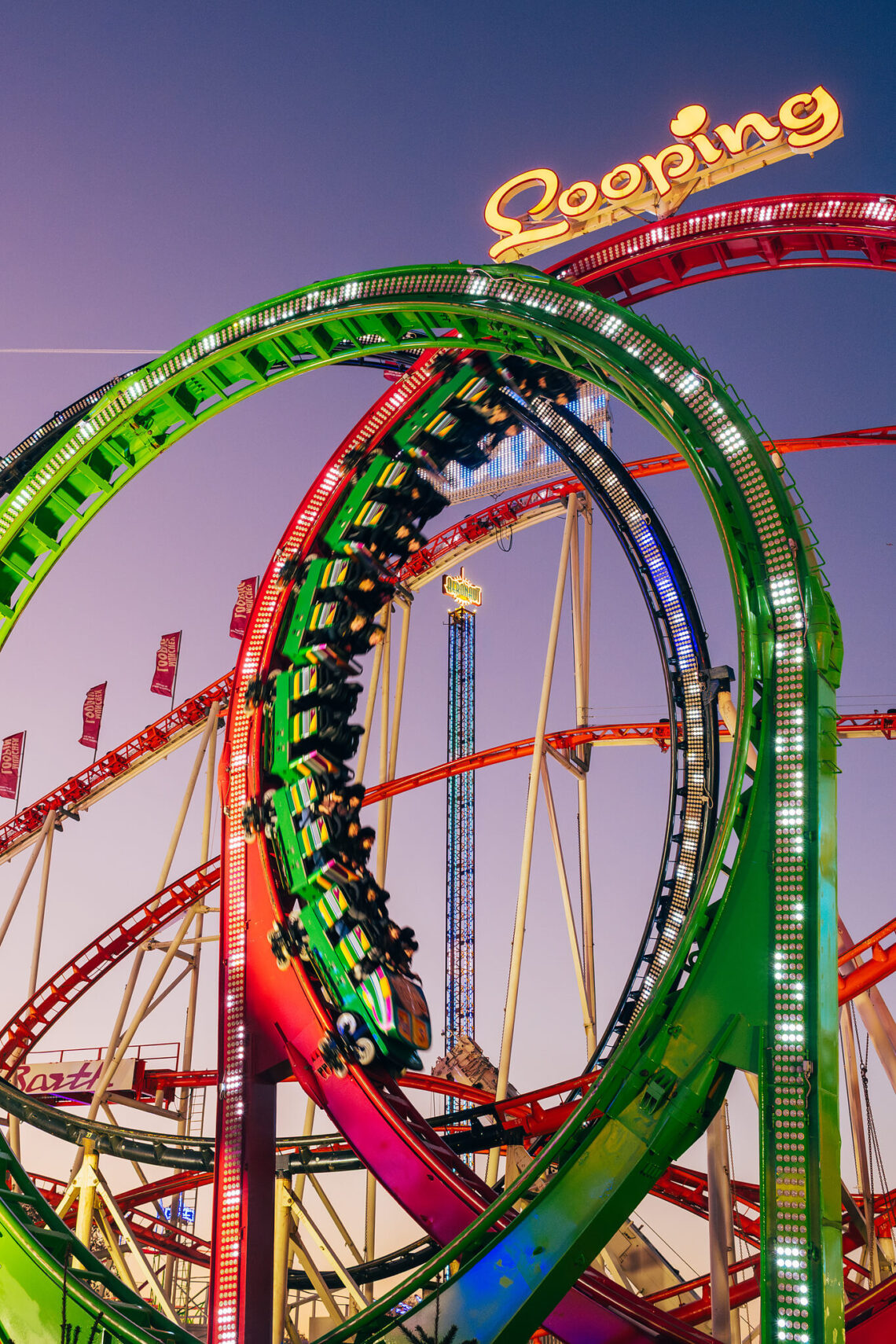 A vibrant roller coaster with neon green and red loops twists against a dusky sky. The track's dramatic loop and lively colors convey excitement.