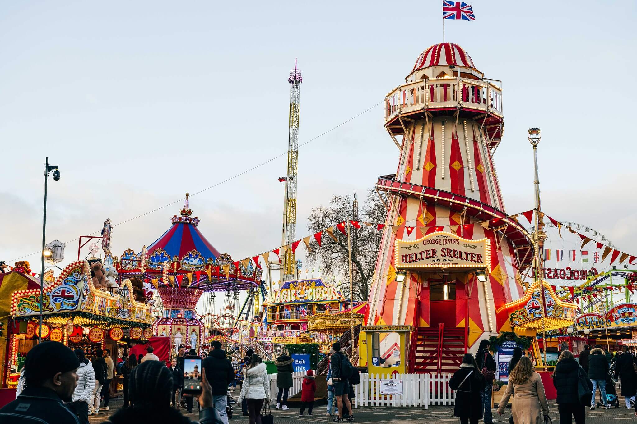 A vibrant fairground scene features a prominent red and yellow striped Helter Skelter ride flying a Union Jack flag on the right. The surrounding amusement park is filled with various colorful attractions, festive bunting, and crowds of visitors exploring the grounds under a pale sky.