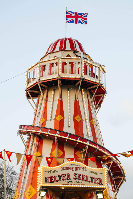 A close up shot features a traditional, lighthouse style Helter Skelter fairground ride decorated with red and yellow vertical stripes and illuminated by rows of warm white lightbulbs. At the top of the tower, a Union Jack flag flies against a clear, pale sky, while a lit sign reading "George Irvin's Helter Skelter" marks the entrance below.