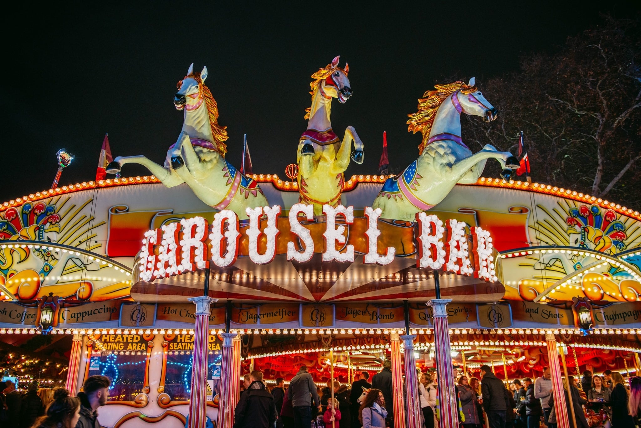 Illuminated Carousel Bar at night with two large, colourful horses on top. Vibrant lights and crowds create a festive, lively atmosphere.