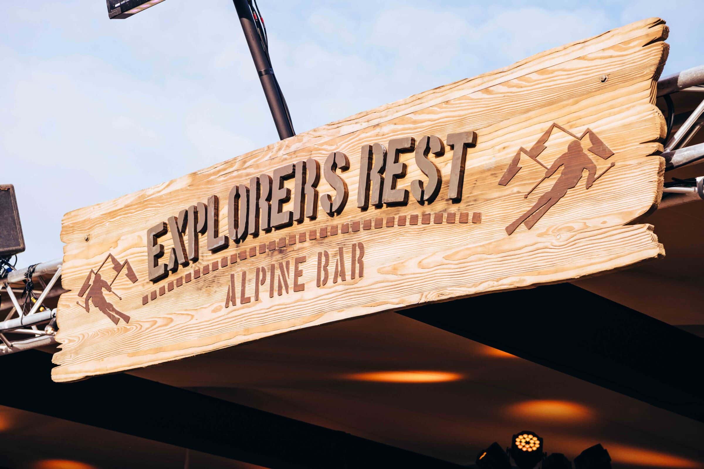 Wooden sign reading "Explorers Rest Alpine Bar" in rustic letters, featuring mountain climbers, under a blue sky. Conveys adventure and relaxation.