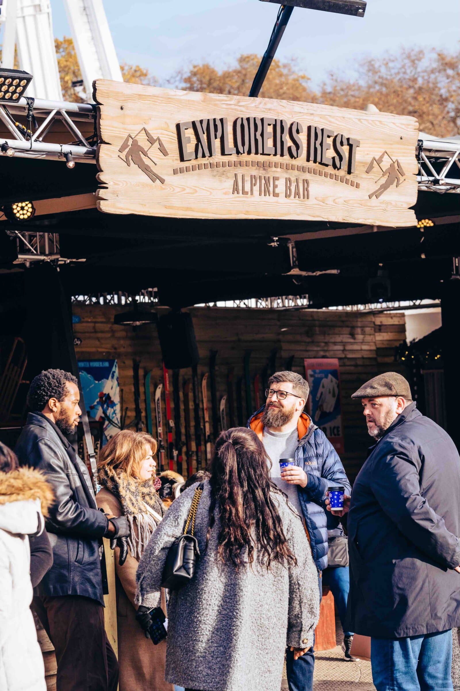 Group of people in winter clothing gather under a wooden sign for the "Explorers Rest Alpine Bar," with skis and winter decor in the background.