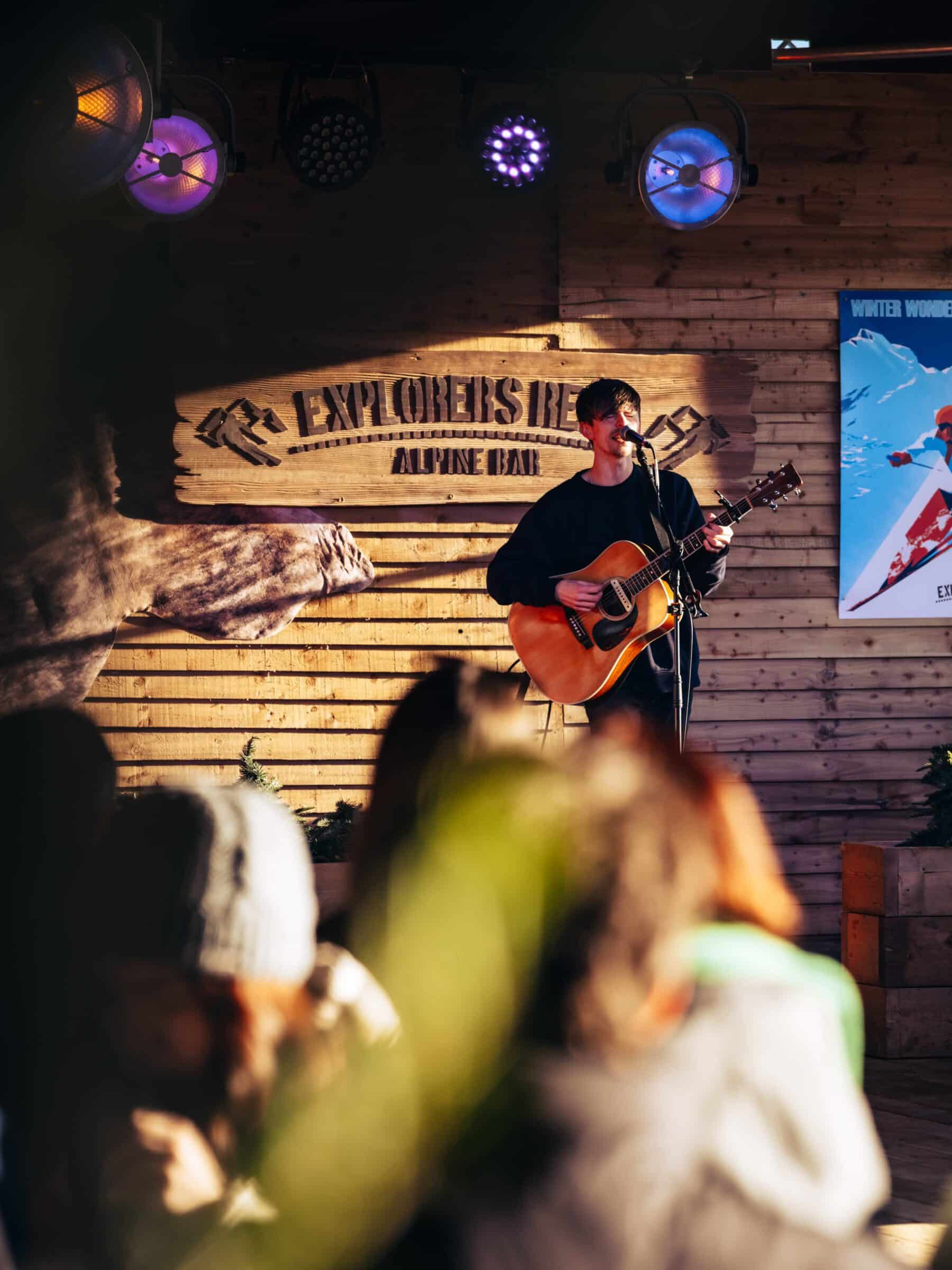 Musician with a guitar performs on stage at Explorers Rest. Warm lighting creates a cozy atmosphere, with an abstract audience in the foreground.