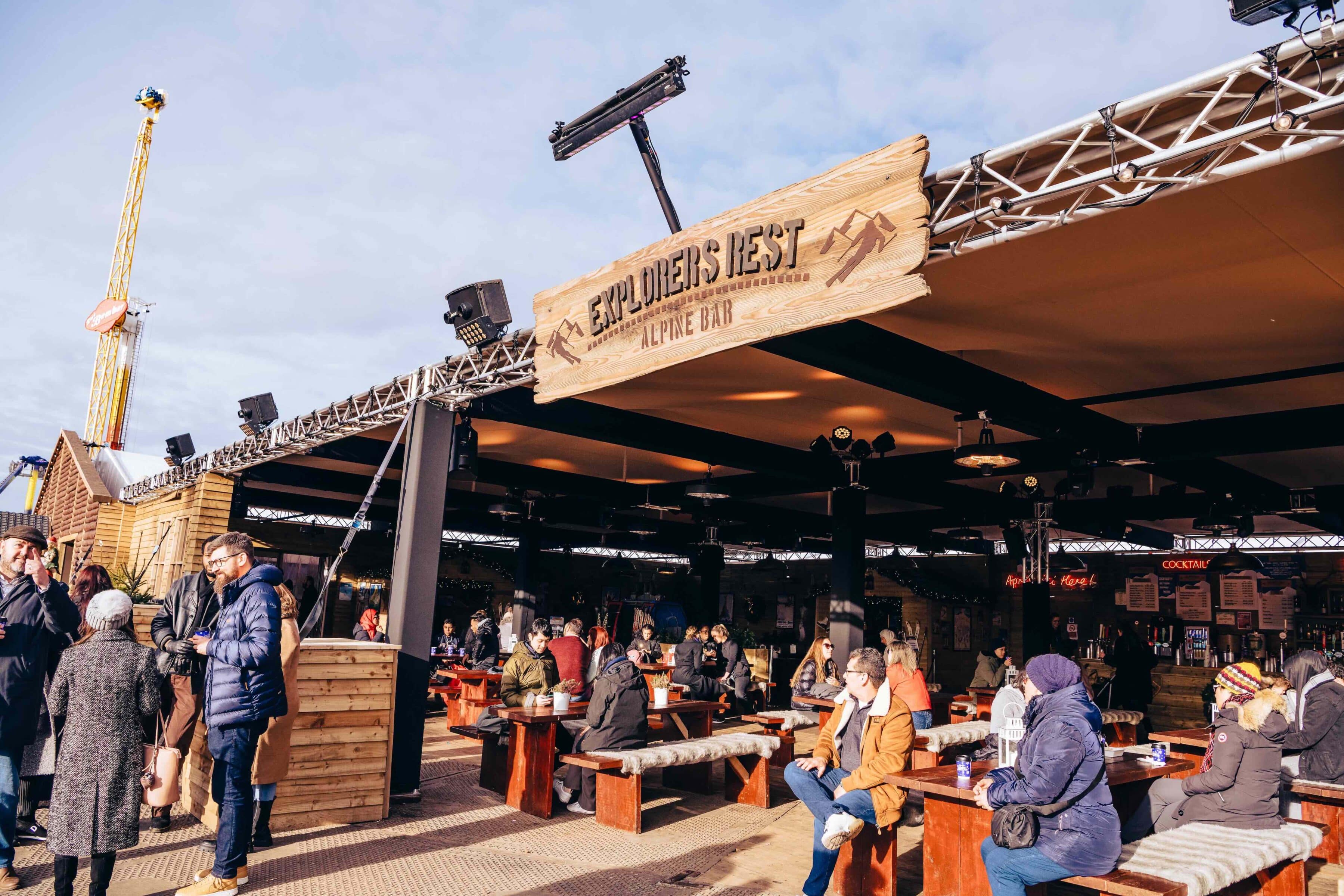 Outdoor alpine bar with a wooden sign reading "Explorers Rest." People in winter clothing sit and chat at wooden tables under a canopy, enjoying a lively atmosphere.