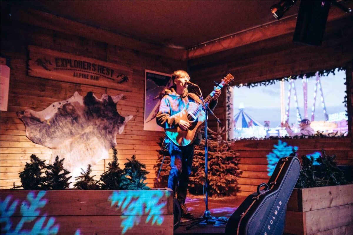 A musician plays guitar and sings on a cozy wooden stage at "Explorers Rest Alpine Bar," surrounded by small pine trees. The scene feels warm and intimate.