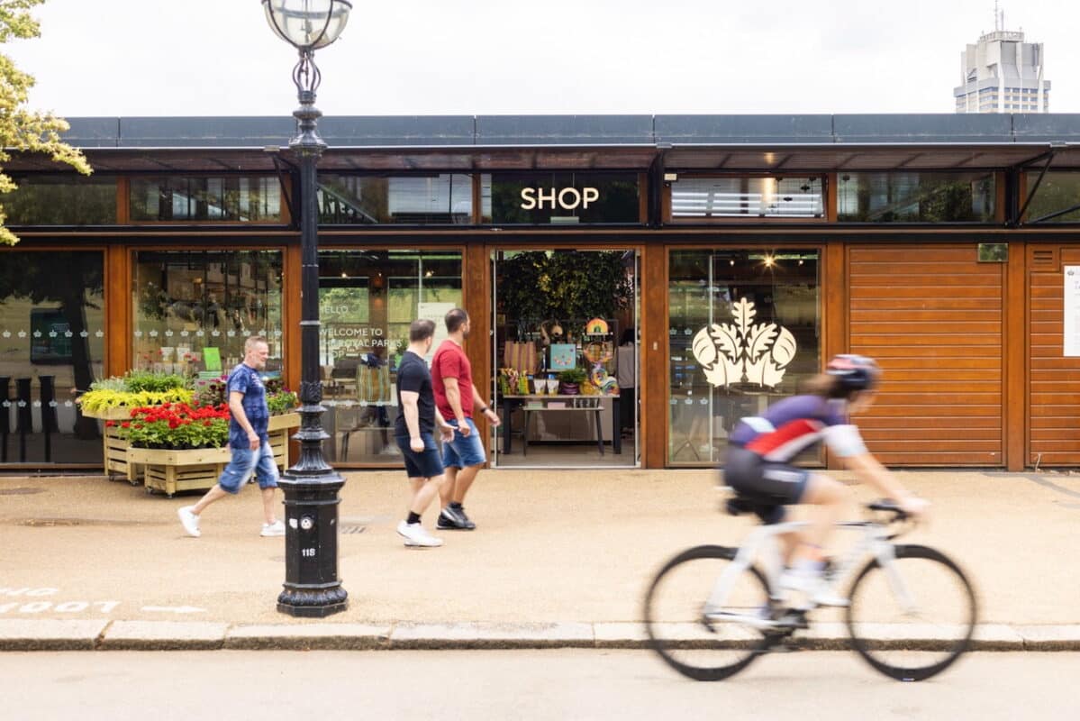 A cyclist in motion passes the Royal Parks' shop exterior with glass doors, people walking by, and vibrant flower displays, creating a lively, busy street scene.