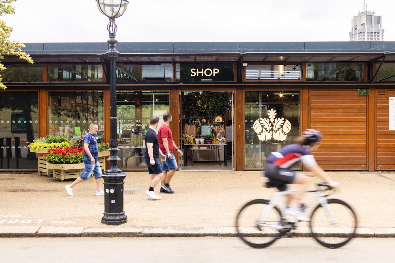 A cyclist in motion passes the Royal Parks' shop exterior with glass doors, people walking by, and vibrant flower displays, creating a lively, busy street scene.