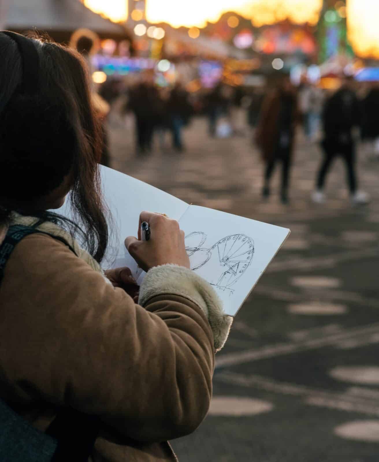 A person sketching a Ferris wheel in a notebook is in focus, with a bustling, blurred crowd and vibrant lights in the background, creating a lively, creative scene.