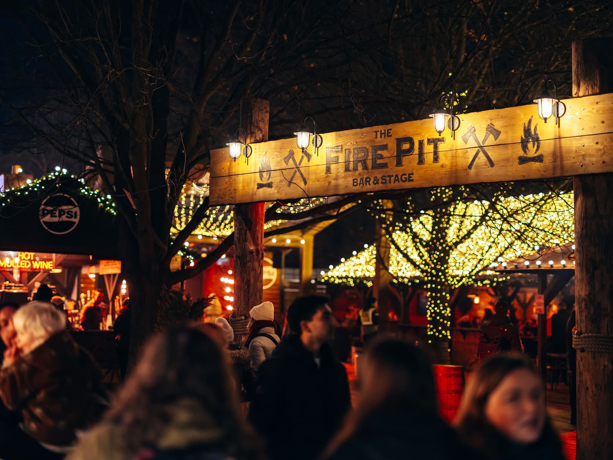 Festive outdoor market at night; illuminated "The Fire Pit Bare & Stage" sign, warm lights, and a crowd enjoying food and drinks. Cozy, cheerful atmosphere.