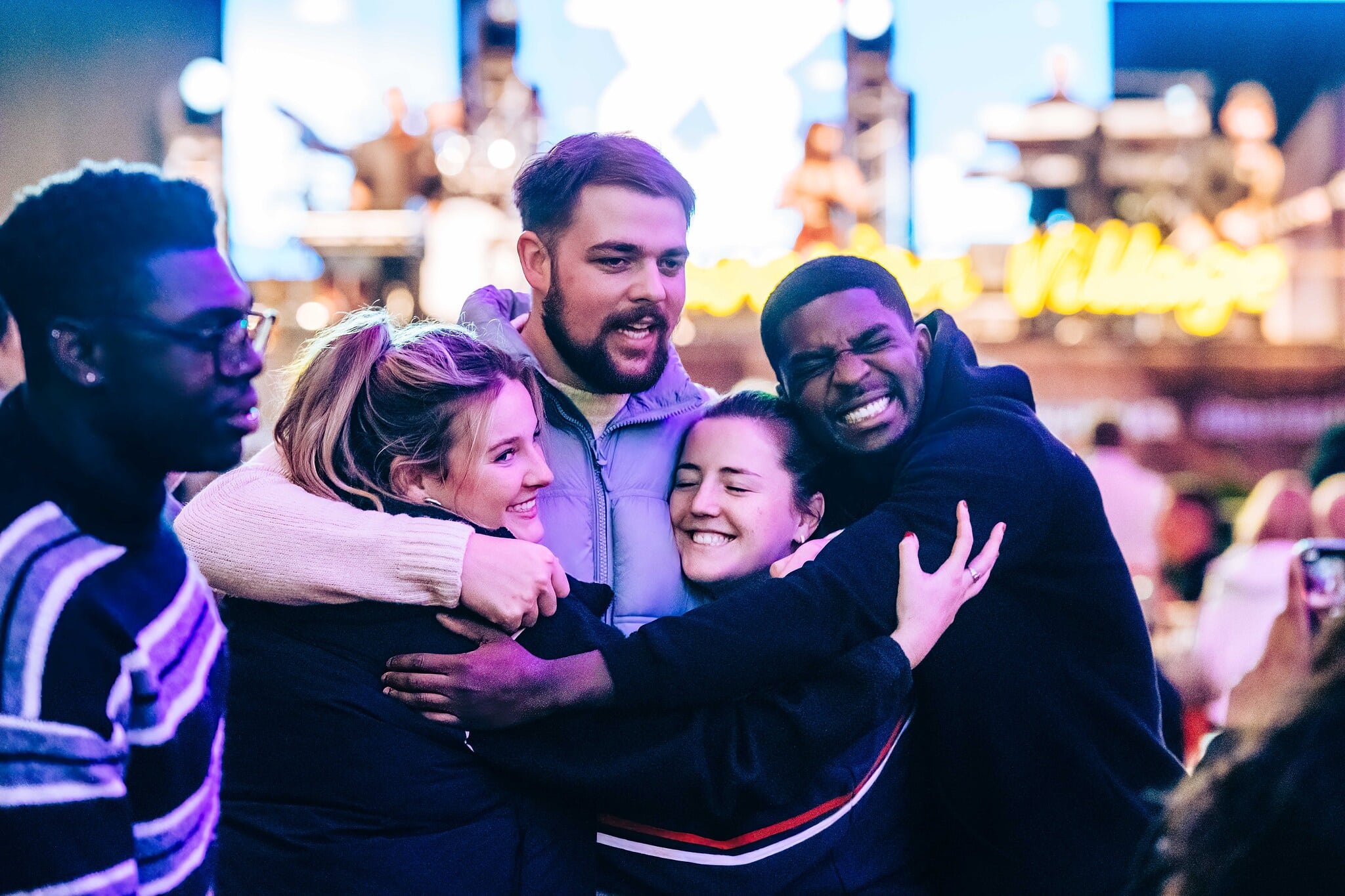 A group of five people share a joyful group hug outdoors at Hyde Park Winter Wonderland. The atmosphere is warm and vibrant, with a stage and colourful lighting in the background.