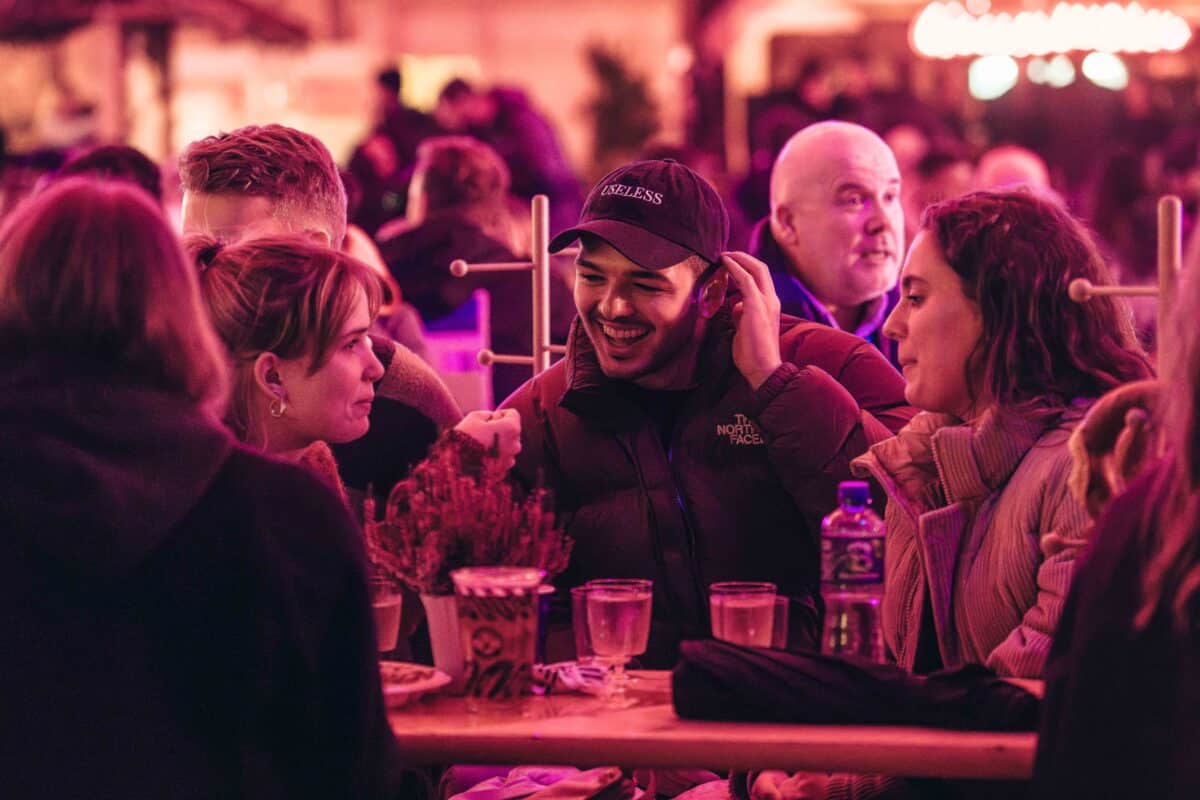 A group of friends sits at a wooden table in a lively, warmly lit setting. They are engaged in conversation and laughter, creating a joyful atmosphere.