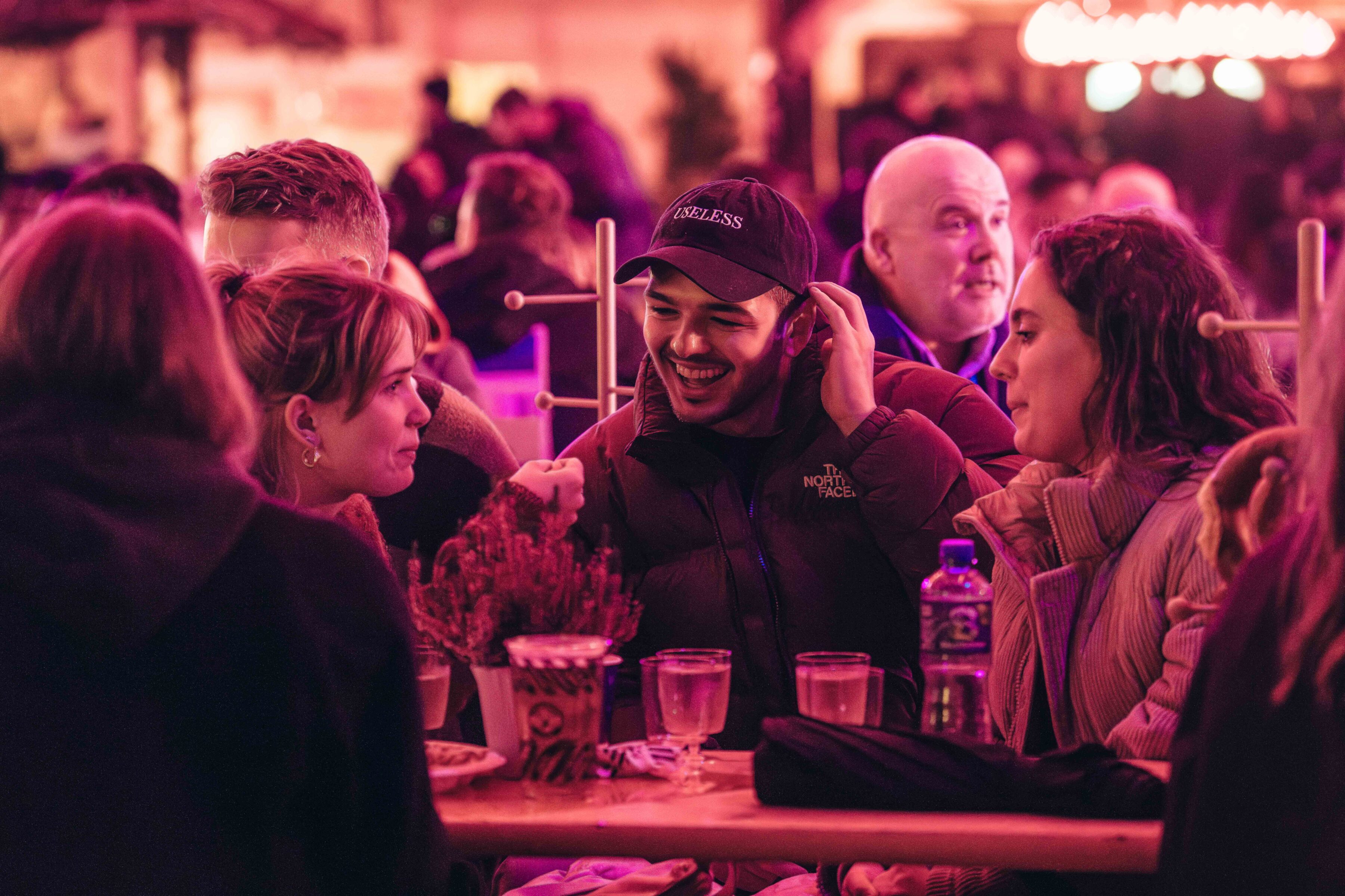 A group of friends sits at a wooden table in a lively, warmly lit setting. They are engaged in conversation and laughter, creating a joyful atmosphere.