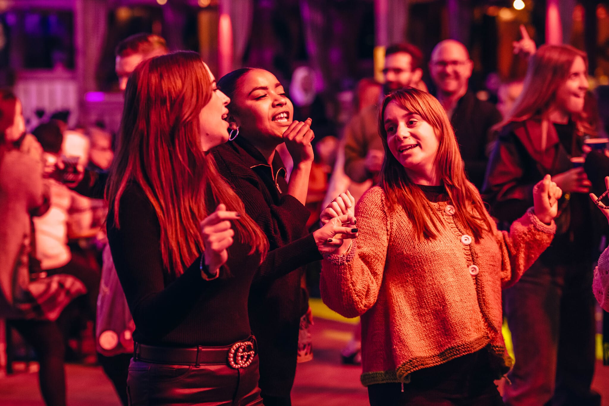 Three people dance joyfully at a lively indoor event at Hyde Park Winter Wonderland, illuminated by warm, colourful lights. Others in the background also appear to be enjoying the celebration.