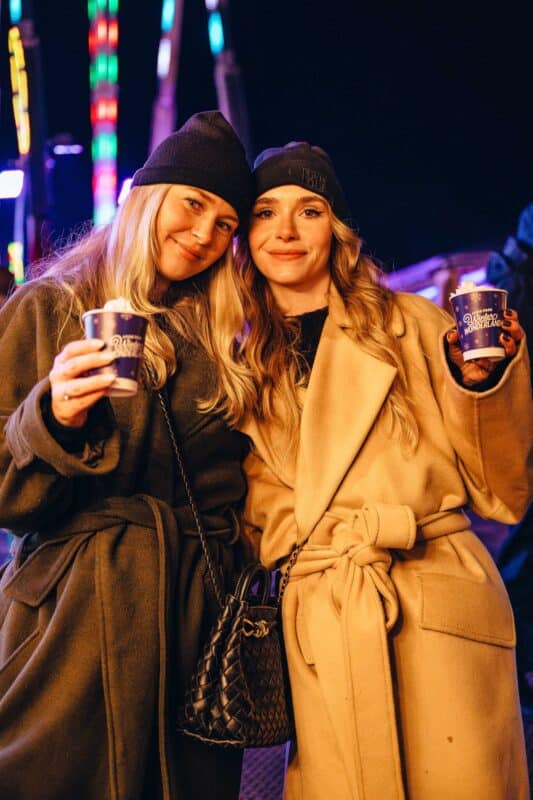 Two women in warm coats and beanies smiling, holding hot drinks in Hyde Park Winter Wonderland branded cups at a festive night event with colourful lights in the background, conveying warmth and joy.