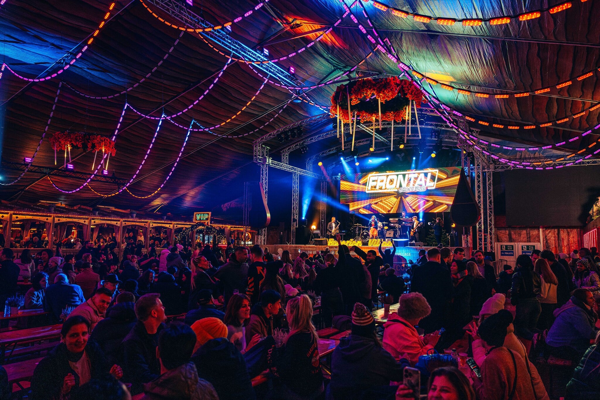 Vibrant festival scene inside a tent at the Bavarian Village with colourful hanging lights and decorations. A lively crowd enjoys a band performing on a well-lit stage.