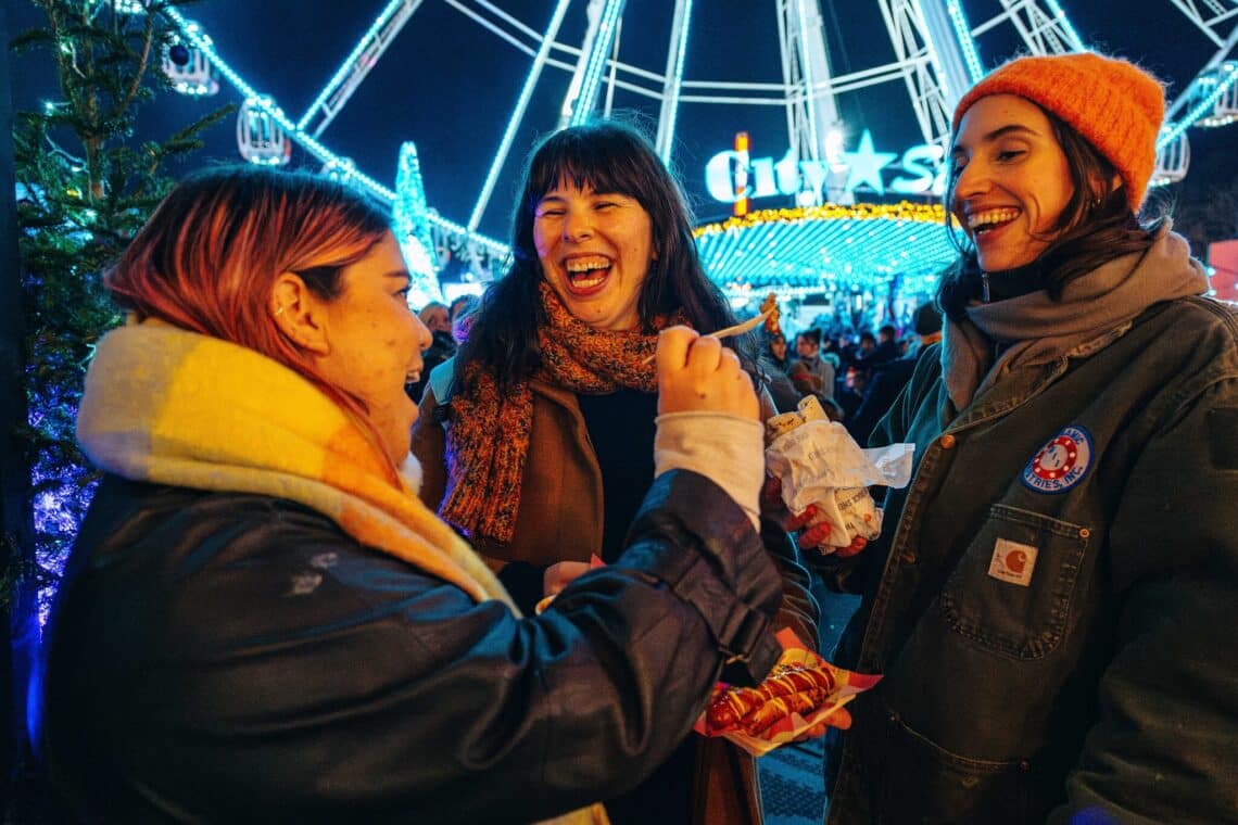 Three friends laughing under festival lights, surrounded by the Giant Wheel presented by Disney+. They wear cozy winter clothes, enjoying a festive atmosphere at night.