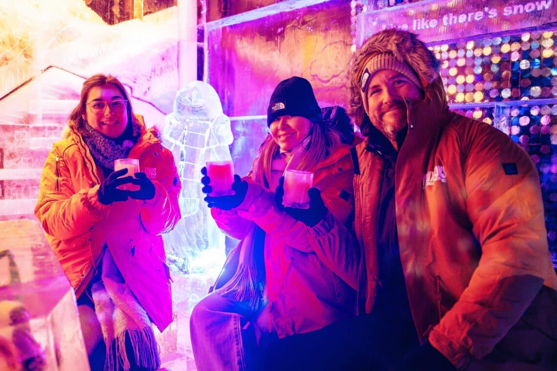 Three people in warm jackets sit in Winter Wonderland's Ice Bar, smiling and holding drinks. Bright purple and orange lights illuminate the ice sculptures around them.