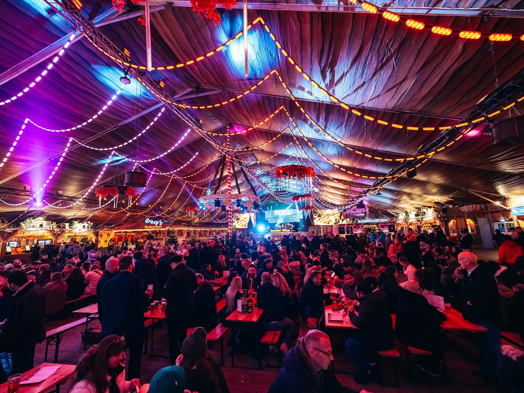 The vibrant Bavarian beer hall at Winter Wonderland with colourful string lights draping from the ceiling, crowded with people at wooden tables, creating a festive, lively atmosphere.