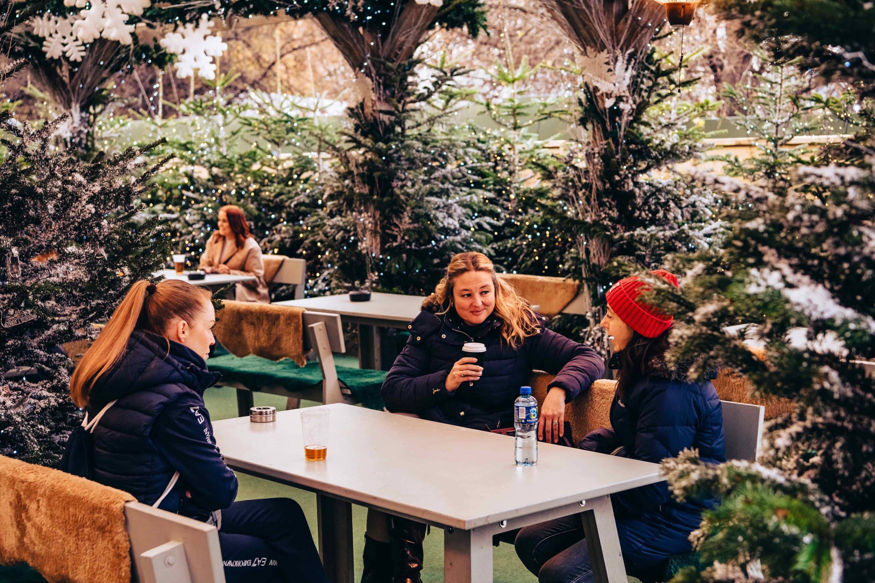 Three women in winter coats chat and drink coffee at Winter Wonderland's Bar Narnia surrounded by snow-dusted evergreens. A cozy, festive atmosphere blooms.