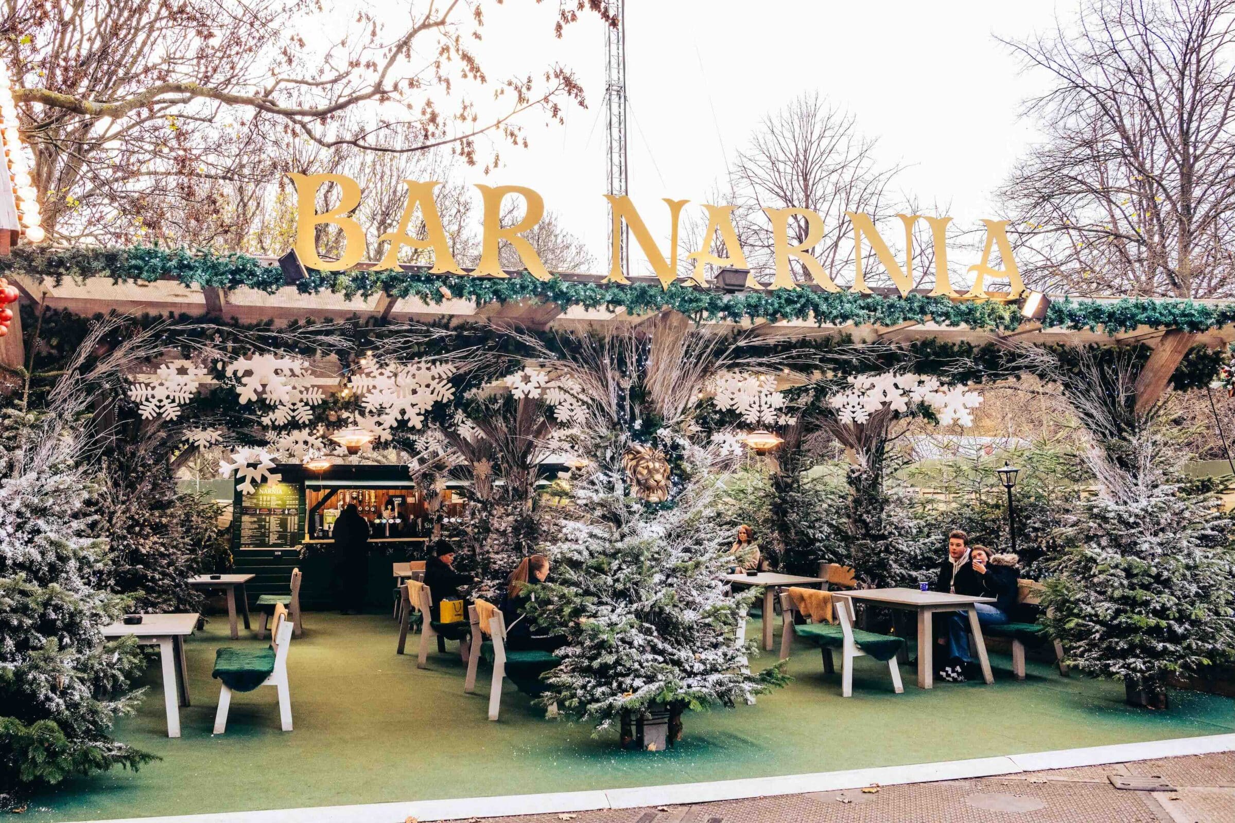Outdoor winter-themed bar named "Bar Narnia" with snowy trees, festive decor, and patrons seated under a canopy, creating a cozy, magical ambiance.