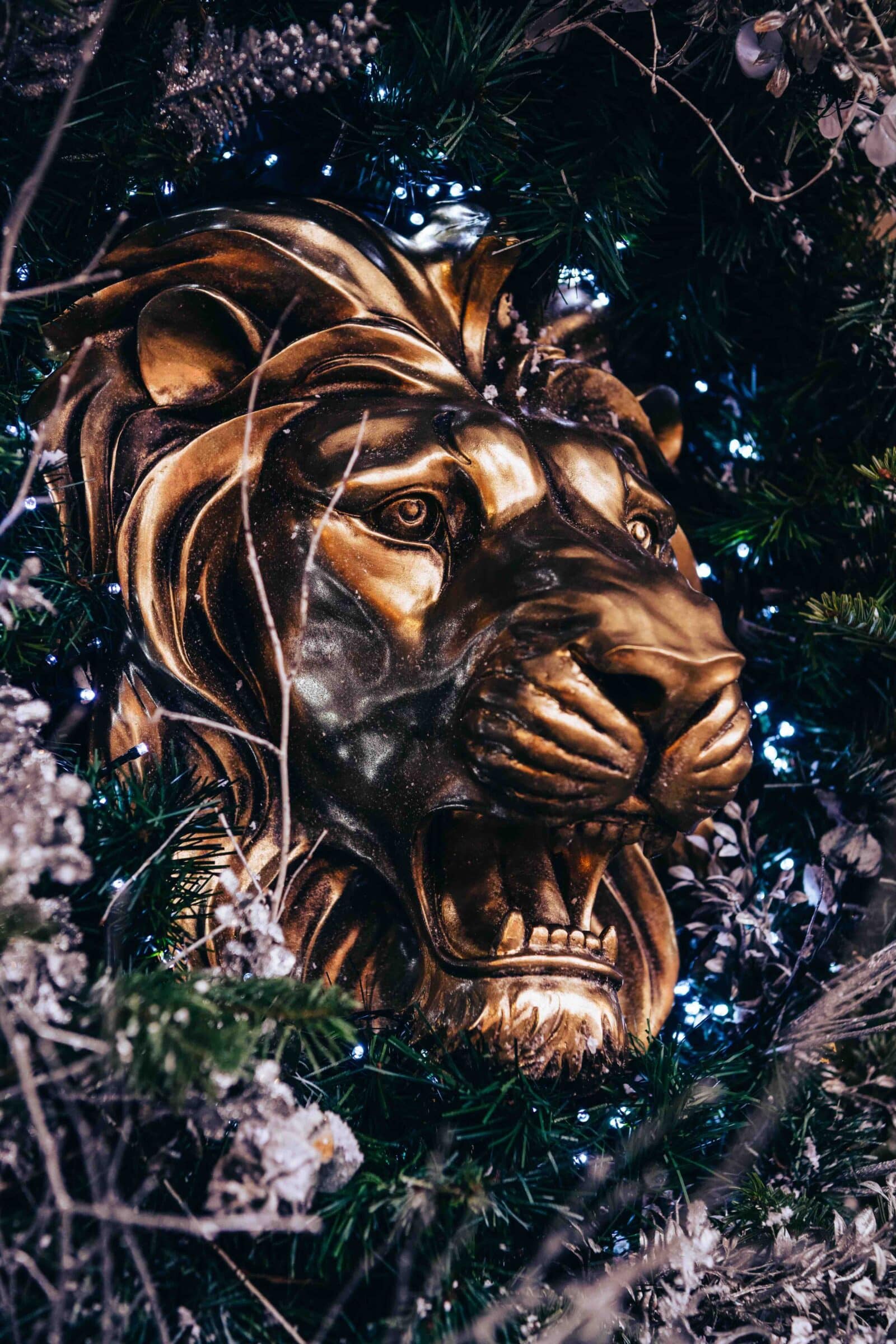Golden lion head sculpture with an open mouth surrounded by green foliage and twinkling lights, evoking a regal and festive atmosphere.