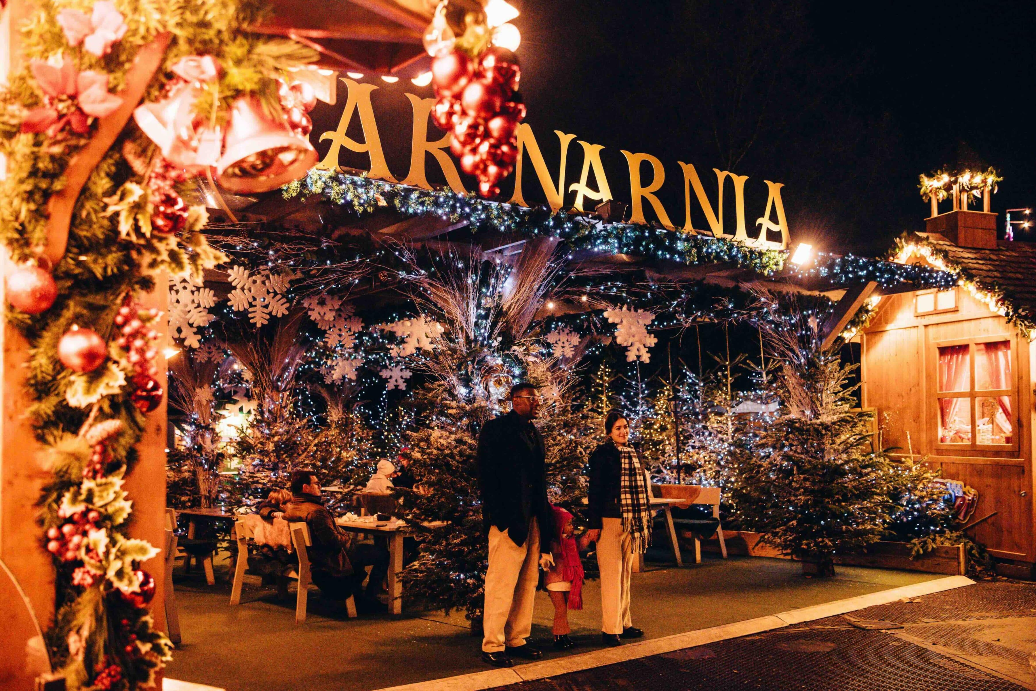 A festive scene at Hyde Park Winter Wonderland's Bar Narnia entrance. Large gold lettering signage, pine trees, snowflakes, and twinkling lights surround visitors, creating a warm and magical atmosphere.