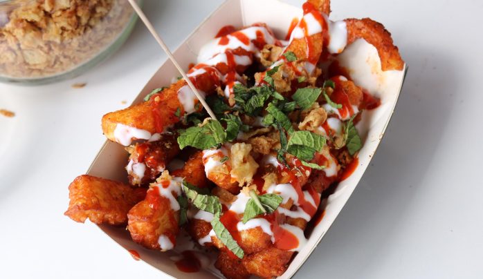 A tray of crispy fried potato wedges drizzled with creamy white sauce and red hot sauce, garnished with fresh green mint leaves and crunchy bits.