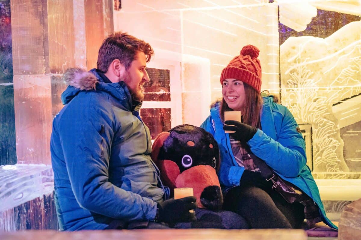 A couple in warm jackets and hats sits on an ice bench, smiling and holding drinks inside Winter Wonderland's Bar Ice. The atmosphere is cozy, with dim, colourful lighting.
