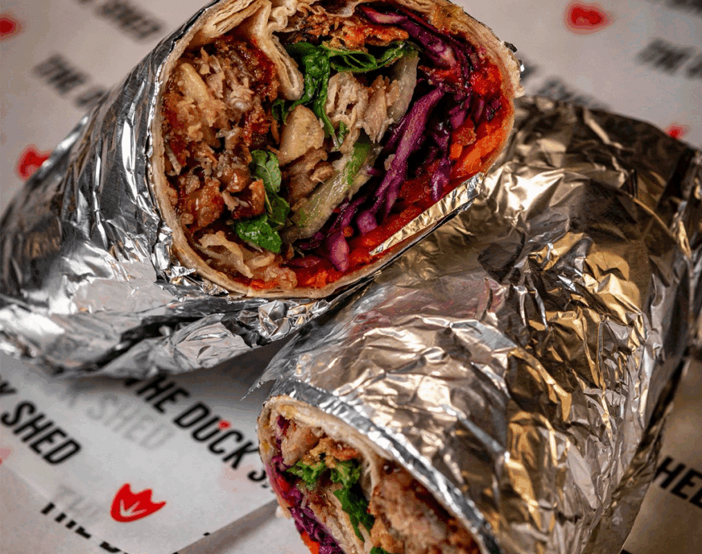 Close-up of two halves of a foil-wrapped burrito, filled with vibrant greens, purple cabbage, orange carrots, and savory meat, on branded paper.