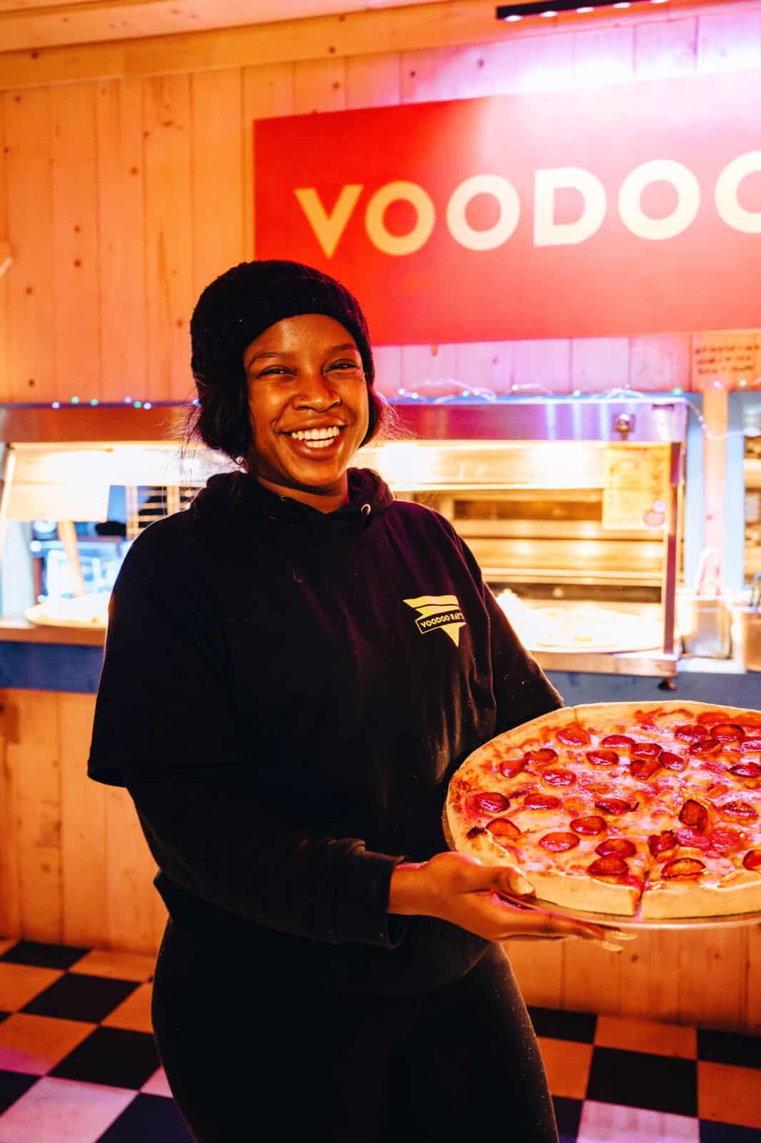 A person joyfully holds a pepperoni pizza in a warmly lit pizza shop with a checkered floor. A bright sign reading "VOODOO" is visible behind them.