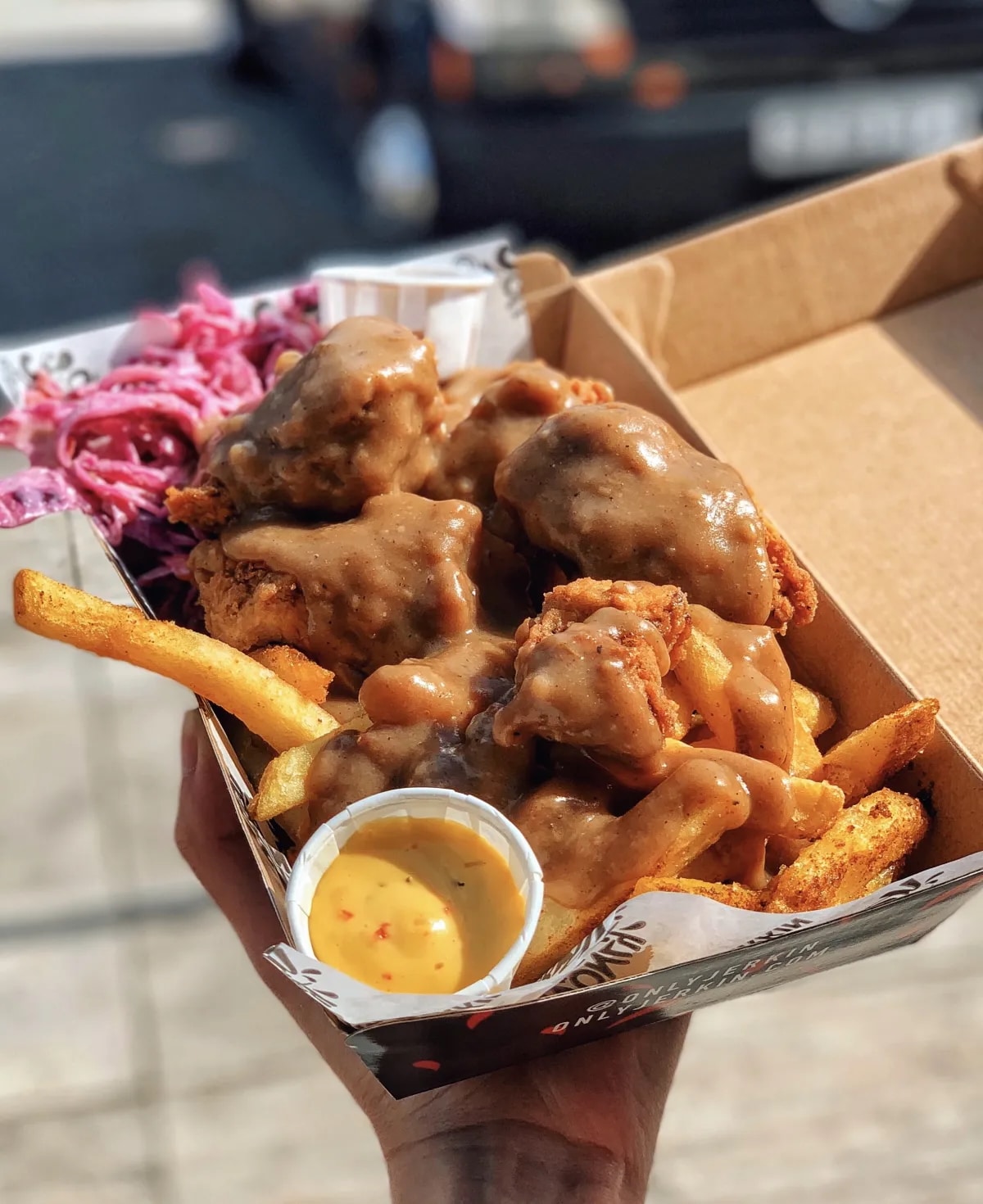 Close-up of a takeout box with crispy fried chicken topped with gravy, golden fries, purple slaw, and a small cup of dipping sauce in sunny light.