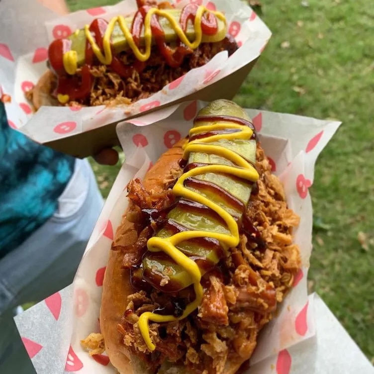 Close-up of two loaded hot dogs in paper trays. Each hot dog is topped with crispy onions, a pickle slice, drizzled with ketchup and mustard. The background shows grass, suggesting an outdoor setting.