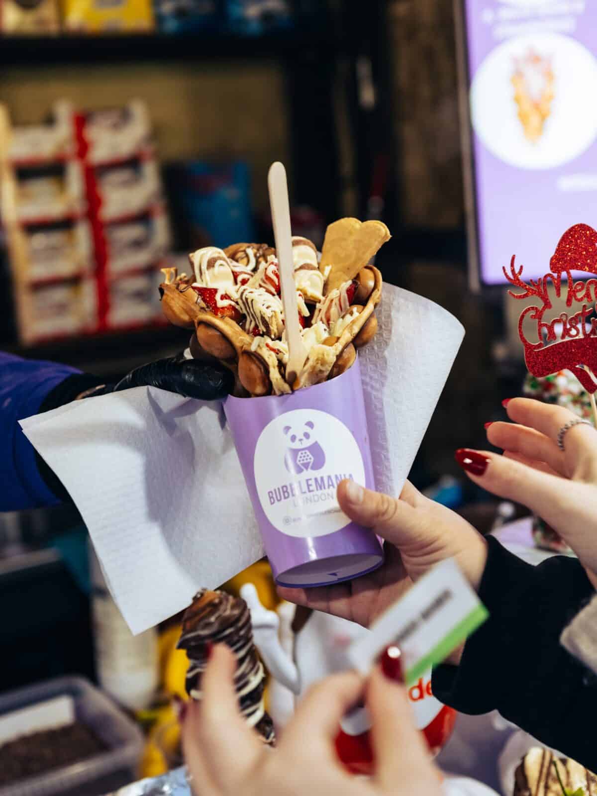 Hands exchange a purple cup holding a bubble waffle with toppings. A red “Merry Christmas” decoration is visible. The scene feels festive and inviting.
