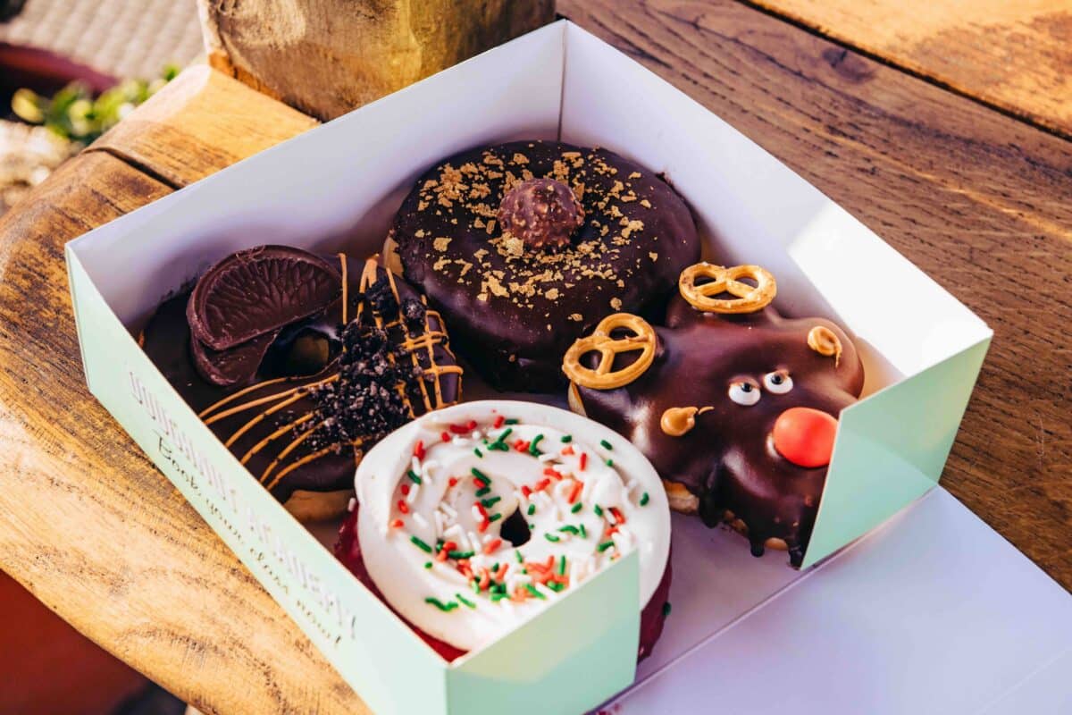 Four assorted, festive donuts in a box on a wooden table. Toppings include chocolate, pretzels, candy eyes, and holiday sprinkles, creating a fun, whimsical vibe.