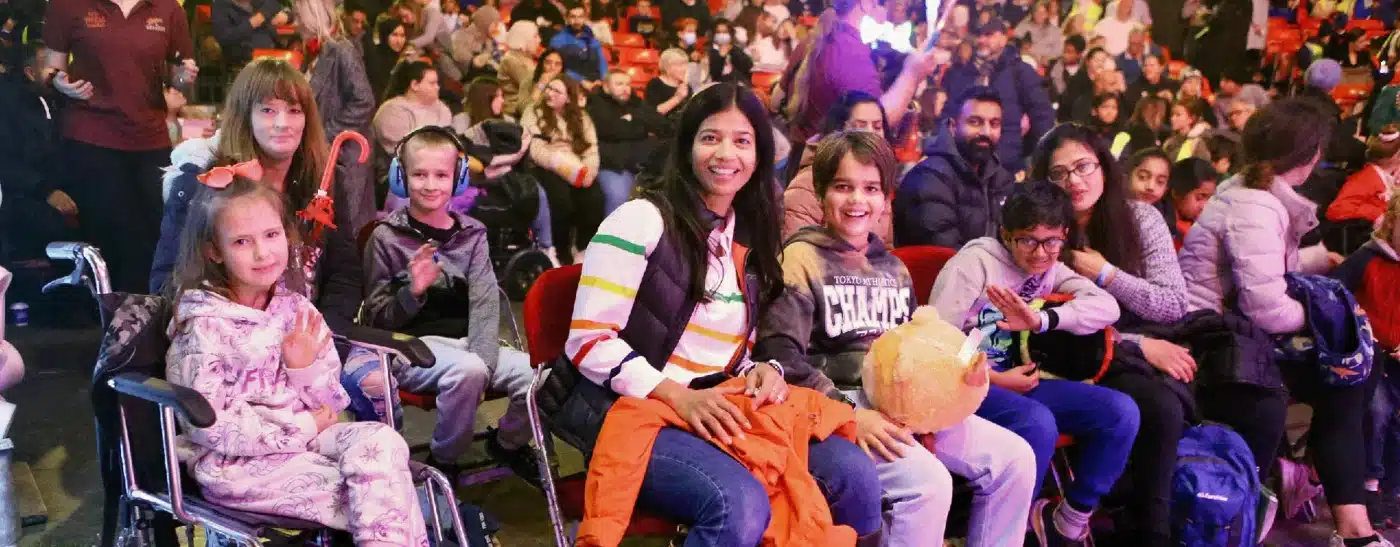 A diverse group of children and adults smile while sitting in a crowded auditorium. A child in a wheelchair appears joyful, creating a warm, inclusive atmosphere.