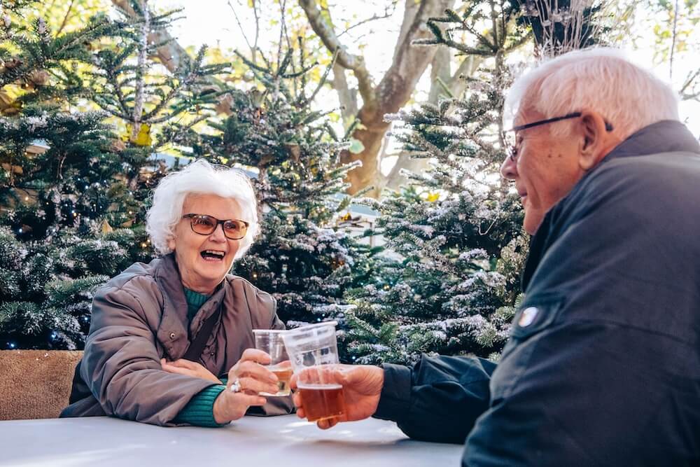 Elderly couple with white hair smiling, toasting with drinks in plastic cups. Seated outdoors, surrounded by frosty pine trees, bright atmosphere.