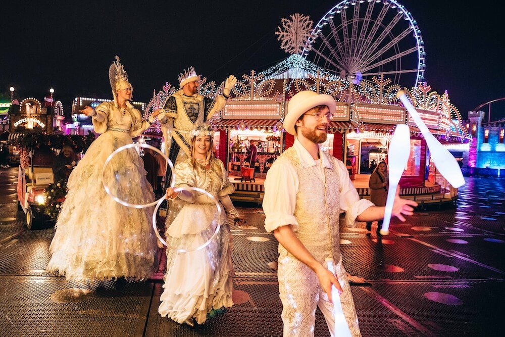 A festive parade with performers in sparkling white costumes, stilt walkers, and a juggler under bright carnival lights. A Ferris wheel glows in the background.