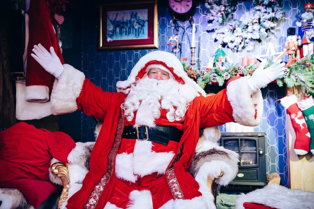 Santa Claus sits joyfully in a festive room with open arms. He's dressed in a classic red suit with white trim. Around him are Christmas decorations including a wreath, stockings, and a vintage television, creating a warm holiday atmosphere.