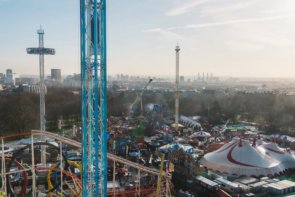 Aerial view of Hyde Park Winter Wonderland with colourful roller coasters and a large circus tent. London city skyline in the background under a clear sky.