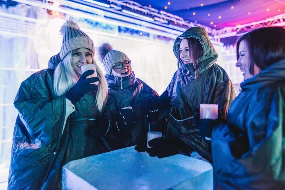 Four people in hooded parkas and beanies smiling and holding drinks in an ice bar. The setting is illuminated with blue and pink lights, creating a cozy atmosphere.
