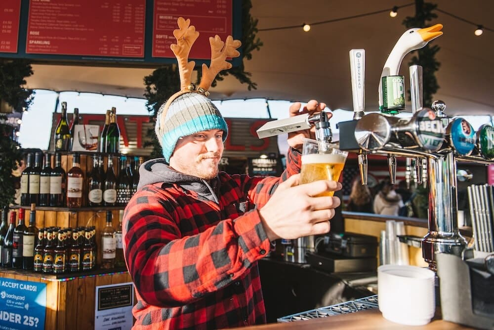 A person in a red plaid shirt and reindeer antlers pours beer from a tap at a festive bar, exuding a cheerful and holiday atmosphere.