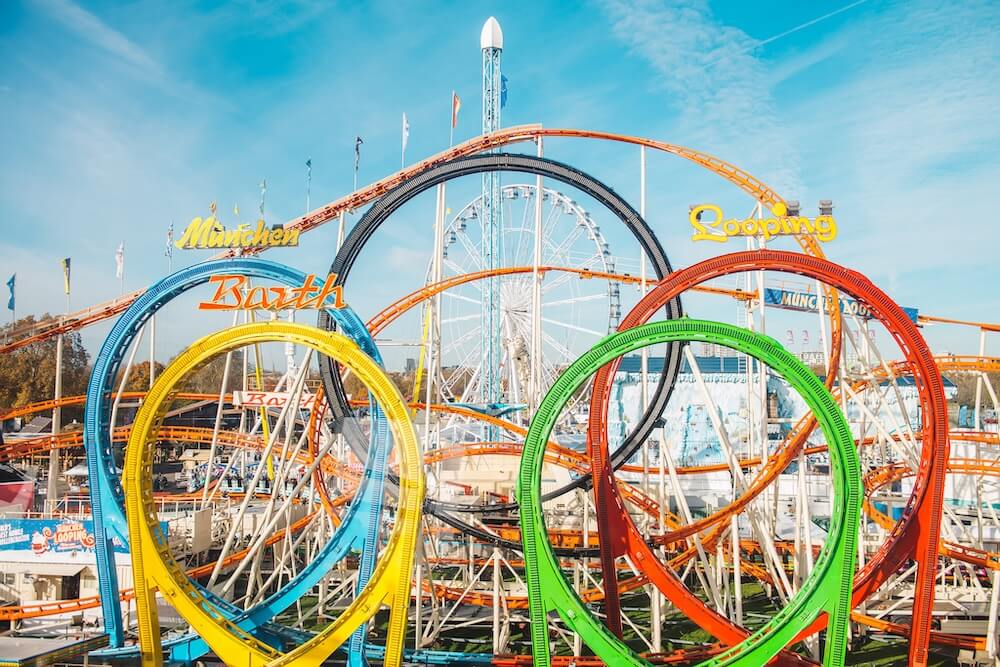 Bright amusement park scene with a u roller coaster featuring five loops in blue, yellow, green, red, and orange, set against a Ferris wheel.