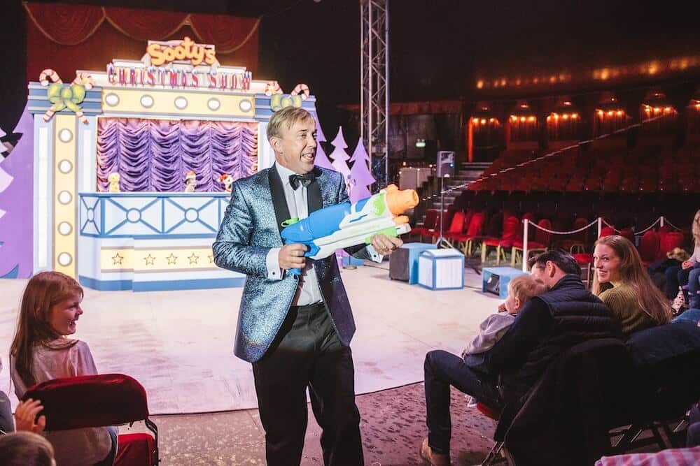 A performer in a sparkling blue jacket holds a toy water gun, smiling at an amused audience during a lively Christmas show in a colourful, festive theatre.