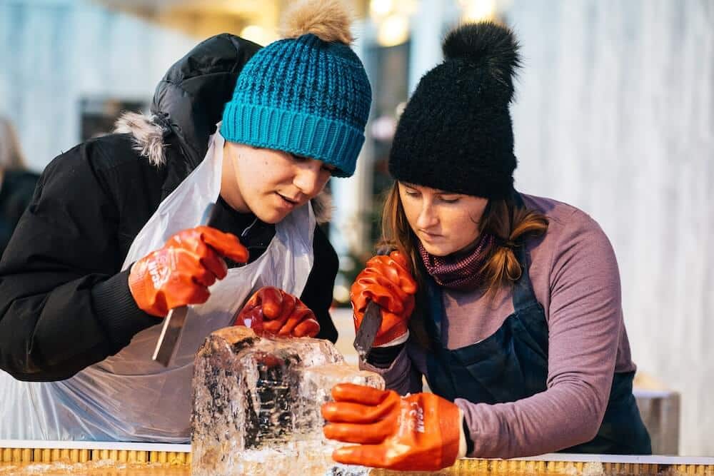 Two people in winter hats and gloves concentrate on carving a large ice block. They use chisels, displaying focus and teamwork in a winter setting.