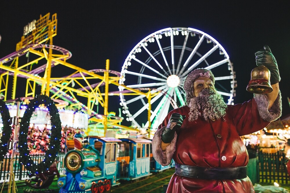 A festive theme park scene at night with a Santa Claus figure holding a bell in the foreground. A brightly lit Ferris wheel and colourful ride tracks are in the background, creating a cheerful and vibrant atmosphere.