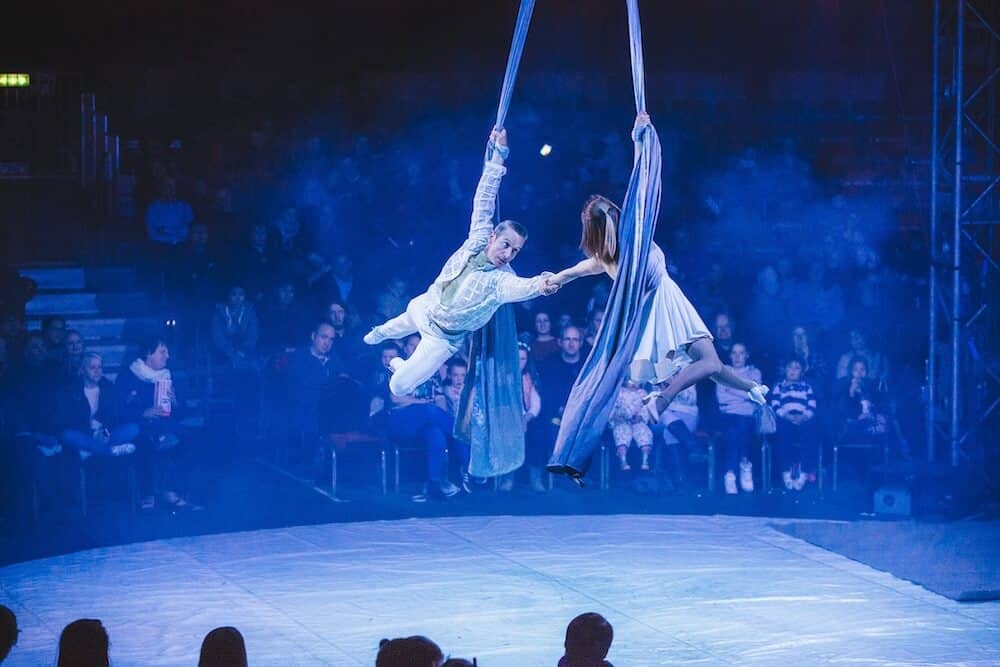Aerial performers in a dimly lit circus hold hands while suspended on blue silks. The audience watches in anticipation, creating a tense and dramatic scene.