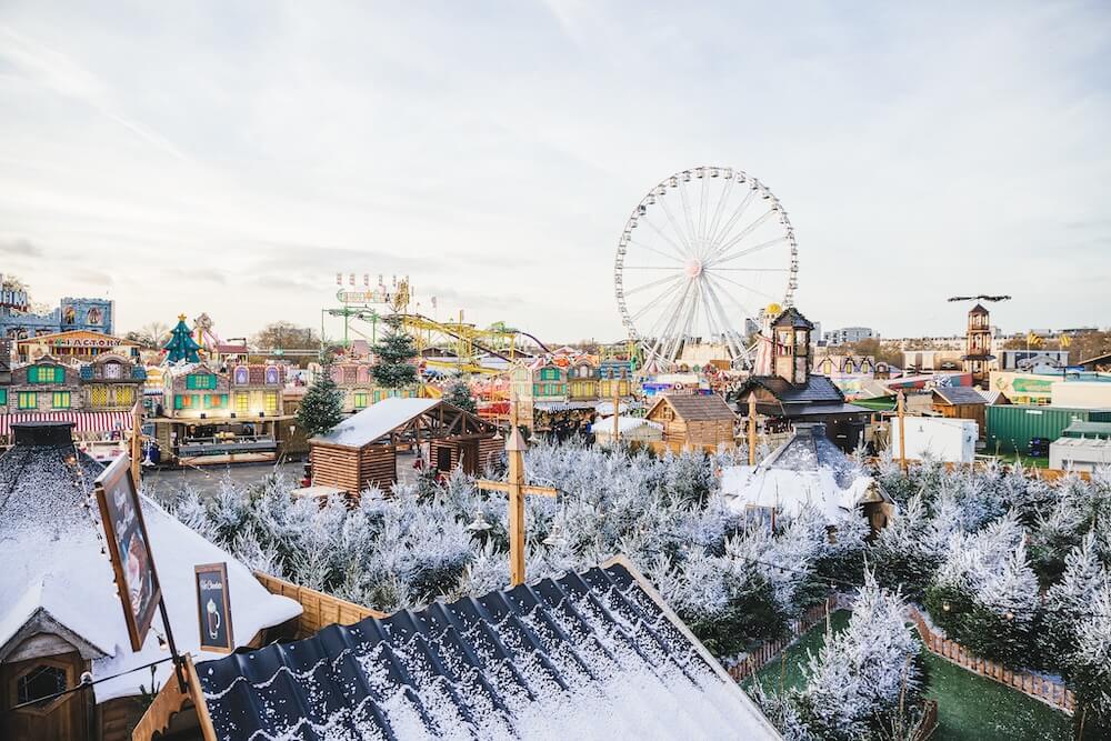 Winter fairground scene with a snow-dusted village, colourful stalls, and evergreen trees. A large Ferris wheel stands in the background under a clear sky.