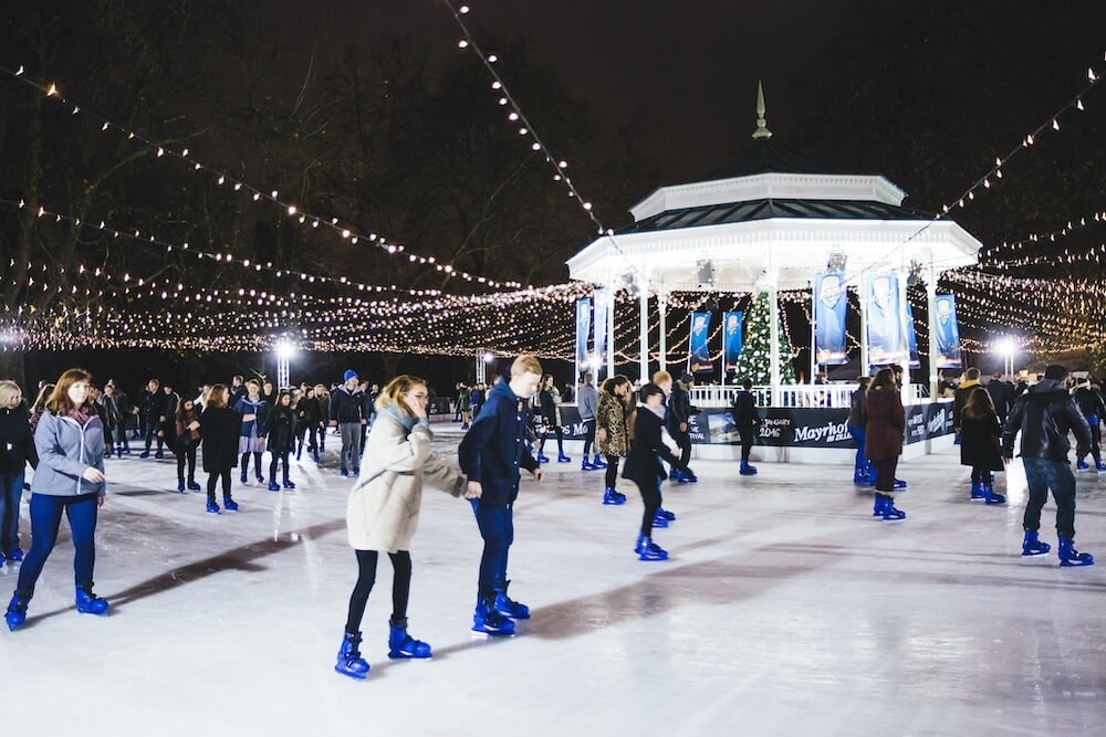 People ice skating on a rink at night, wearing blue skates. A gazebo is decorated with lights and a Christmas tree, creating a festive atmosphere.